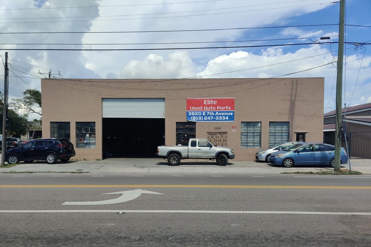 Elite Used Auto: Tan brick auto parts building with a large roll-up door, red and blue sign, and a pickup and cars parked out front