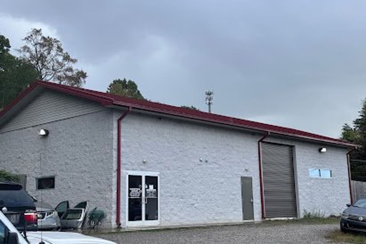 European Import Auto Parts: Gray block building with a red metal roof, double glass entrance door and a roll-up garage door, with cars parked in the gravel lot