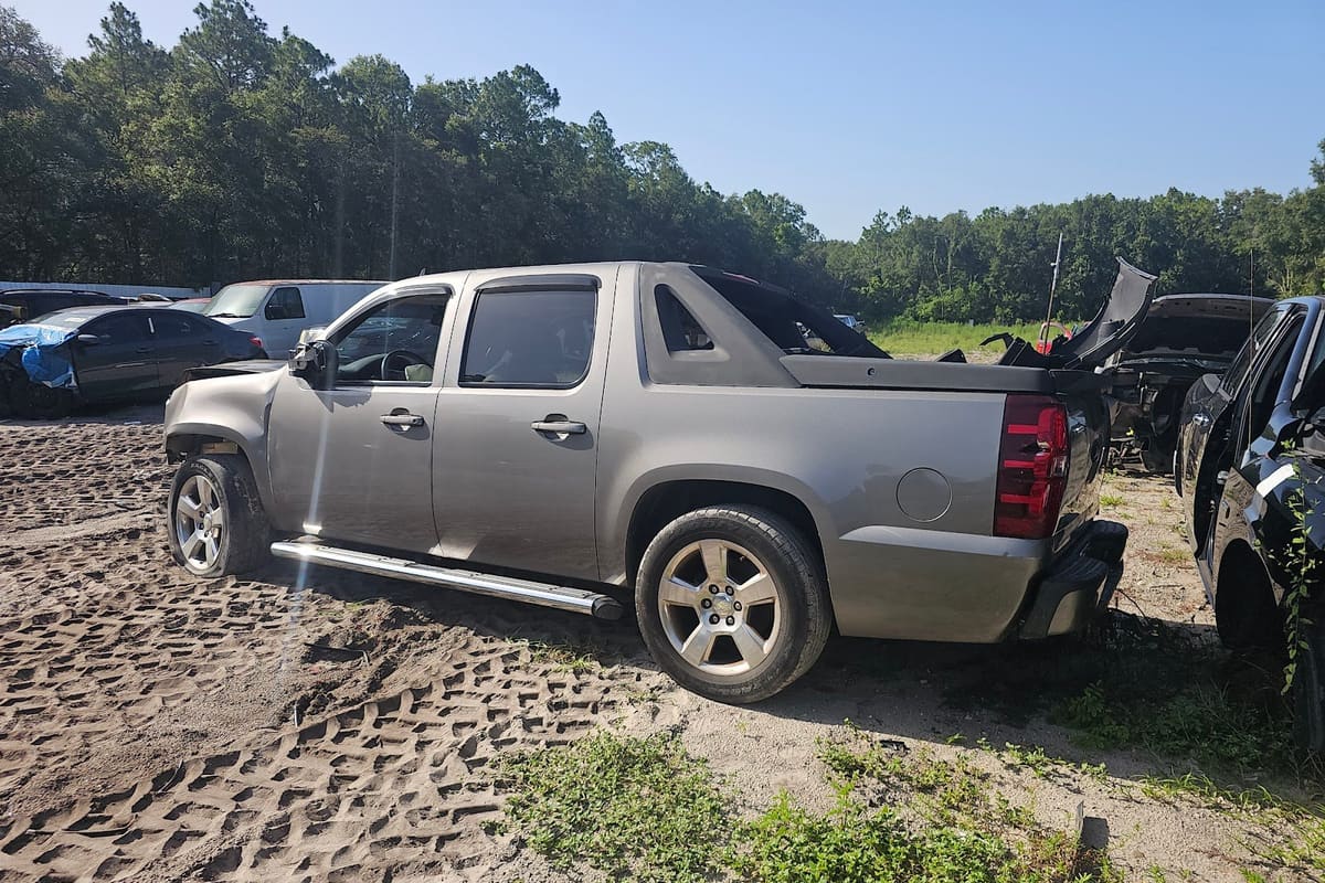 M&M Auto Recycling: Wrecked gray pickup truck parked on a sandy salvage lot with other damaged vehicles and trees behind
