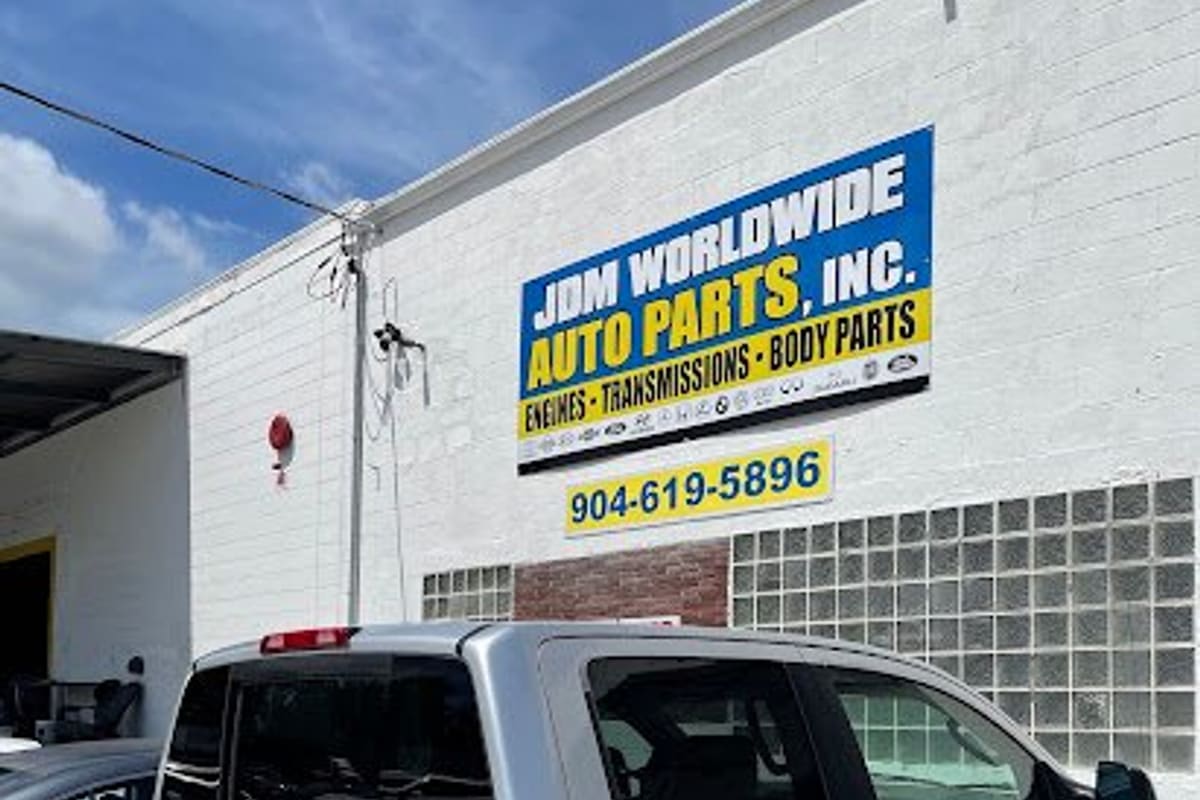JDM Worldwide Auto Parts: White brick building with a blue and yellow auto parts sign and phone number, a silver pickup truck parked in front
