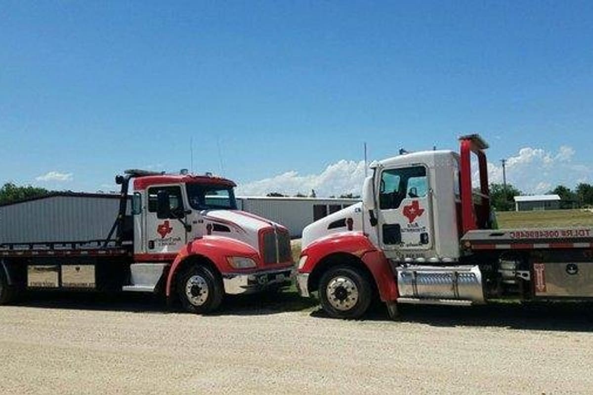Robin Pack: Two red and white flatbed tow trucks parked on a gravel lot with metal buildings under a blue sky