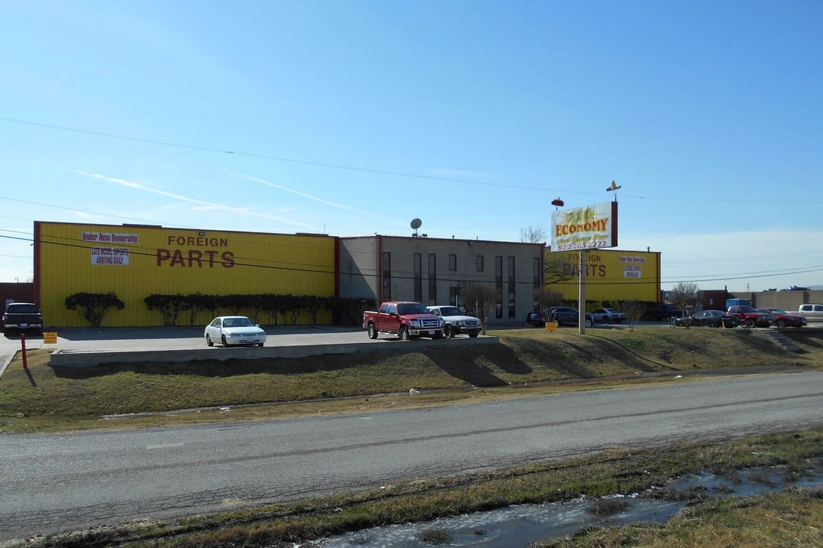 DMR Import Recyclers: Yellow auto parts building with a tall roadside sign and vehicles parked in front under a clear blue sky