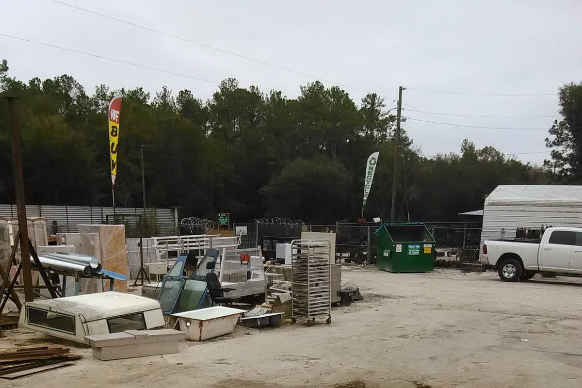 Waltrip Salvage and Scrap: Wide view of a dirt scrap yard with stacked metal, bathtubs, racks and a green dumpster, with a pickup truck at right