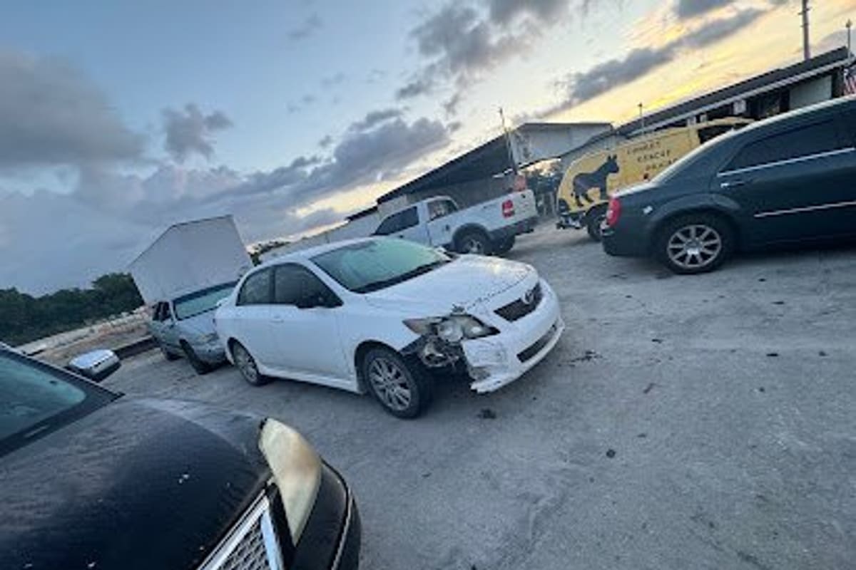 Mims Pick & Pay Recycler Center: Damaged white sedan and other vehicles parked on a gravel lot with low sheds and a pickup truck in the background at dusk