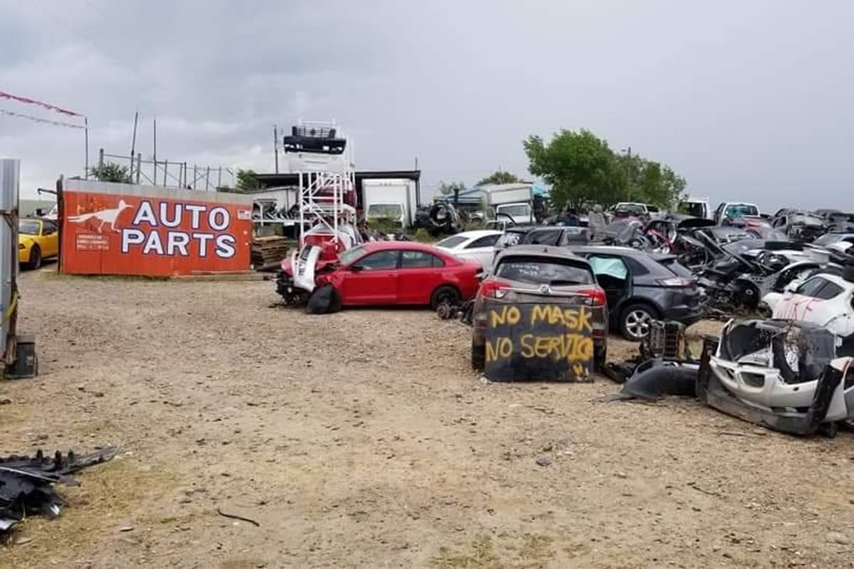 Paisano Used Auto Parts: Wide view of a salvage yard with rows of wrecked cars on gravel and an orange auto parts sign in the background