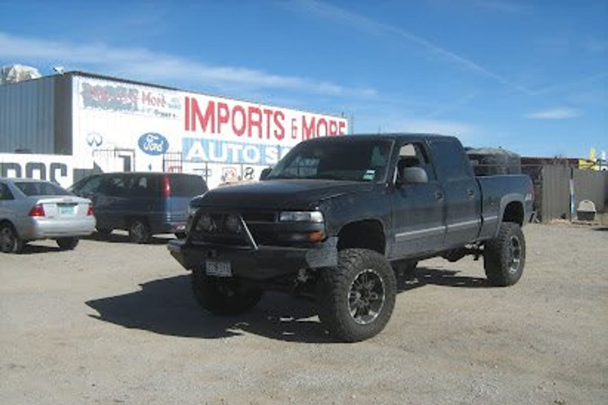 Imports & More Auto Salvage: Large red and white building sign reading Imports & More Auto above a gravel lot with a lifted black pickup and parked cars