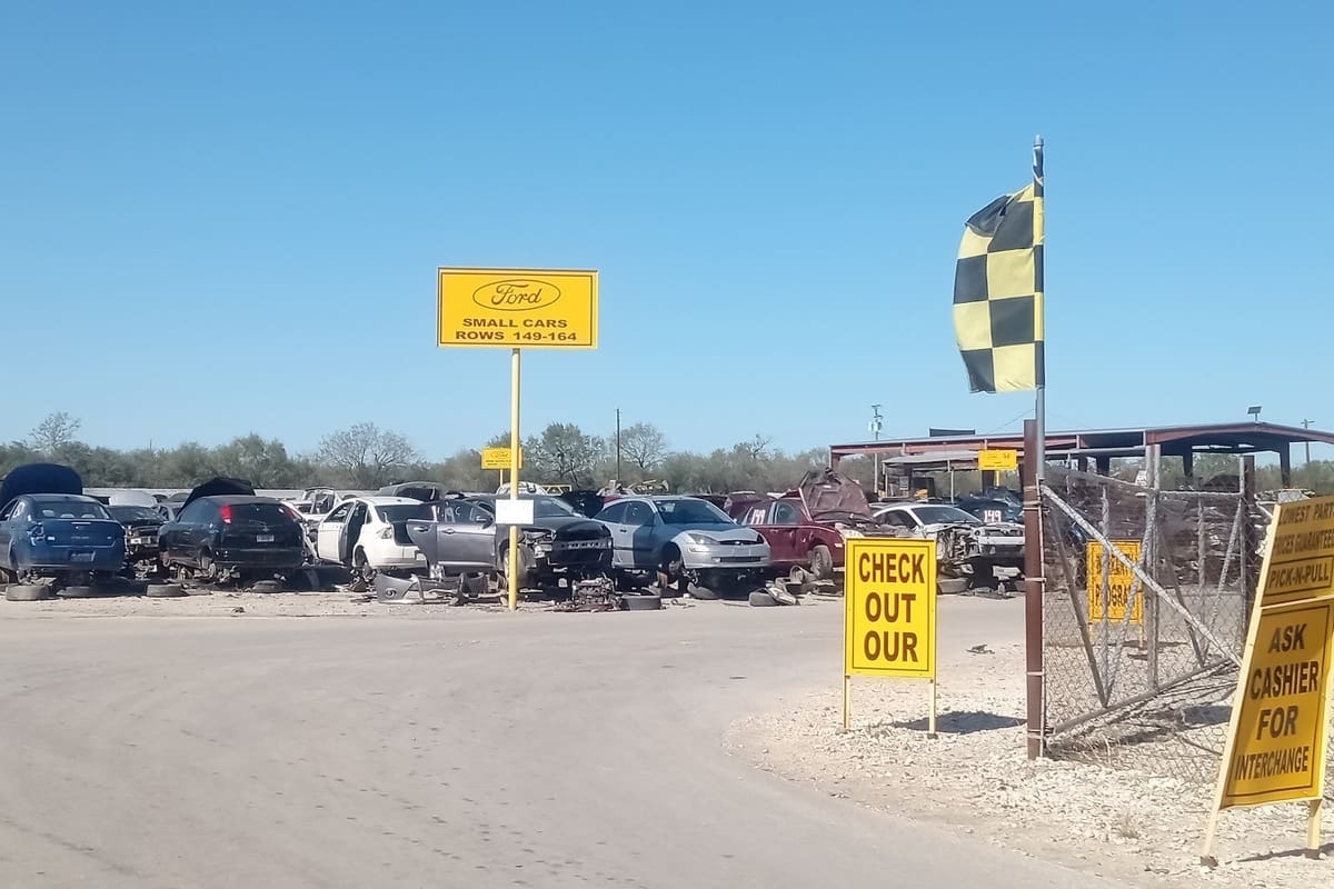 Pick-N-Pull: Wide daytime view of a salvage yard with rows of cars, a yellow row sign, a checkered flag and yellow signs near the entrance