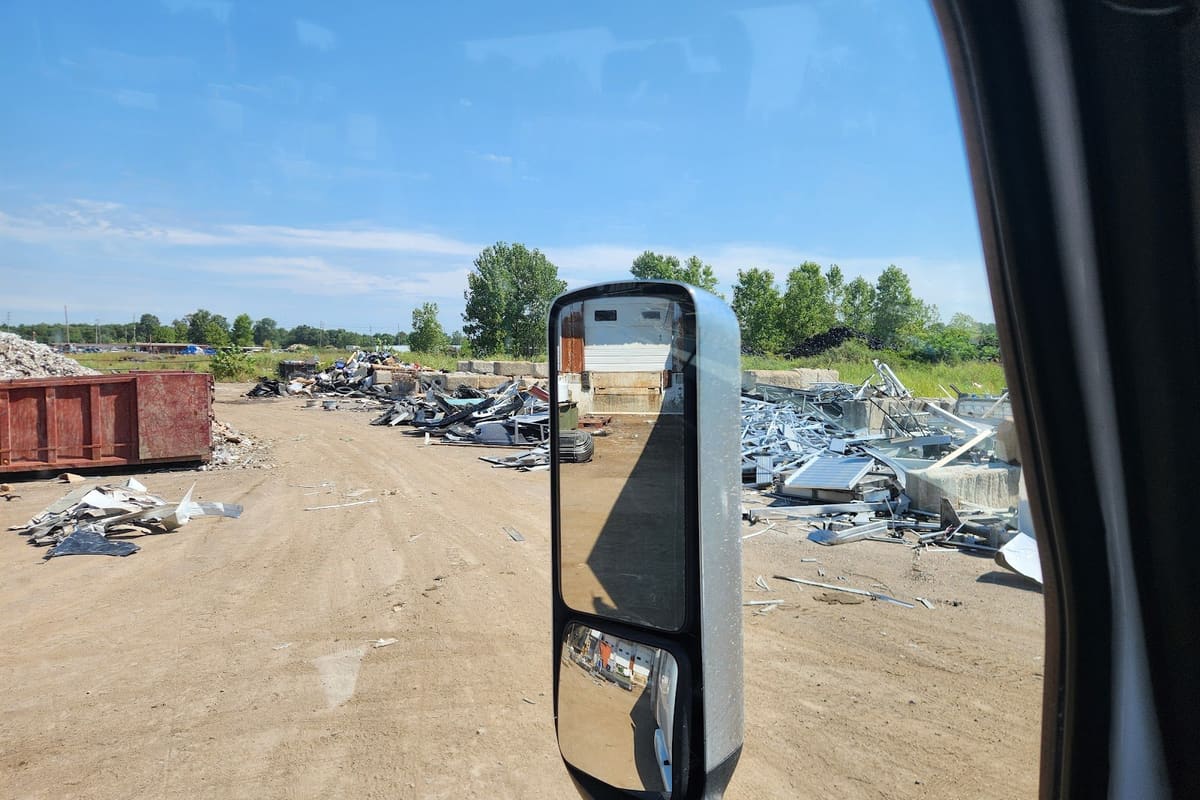 Worley Recyclling: Dirt scrap yard with piles of scrap metal and a red dumpster seen through a truck's side mirror on a sunny day