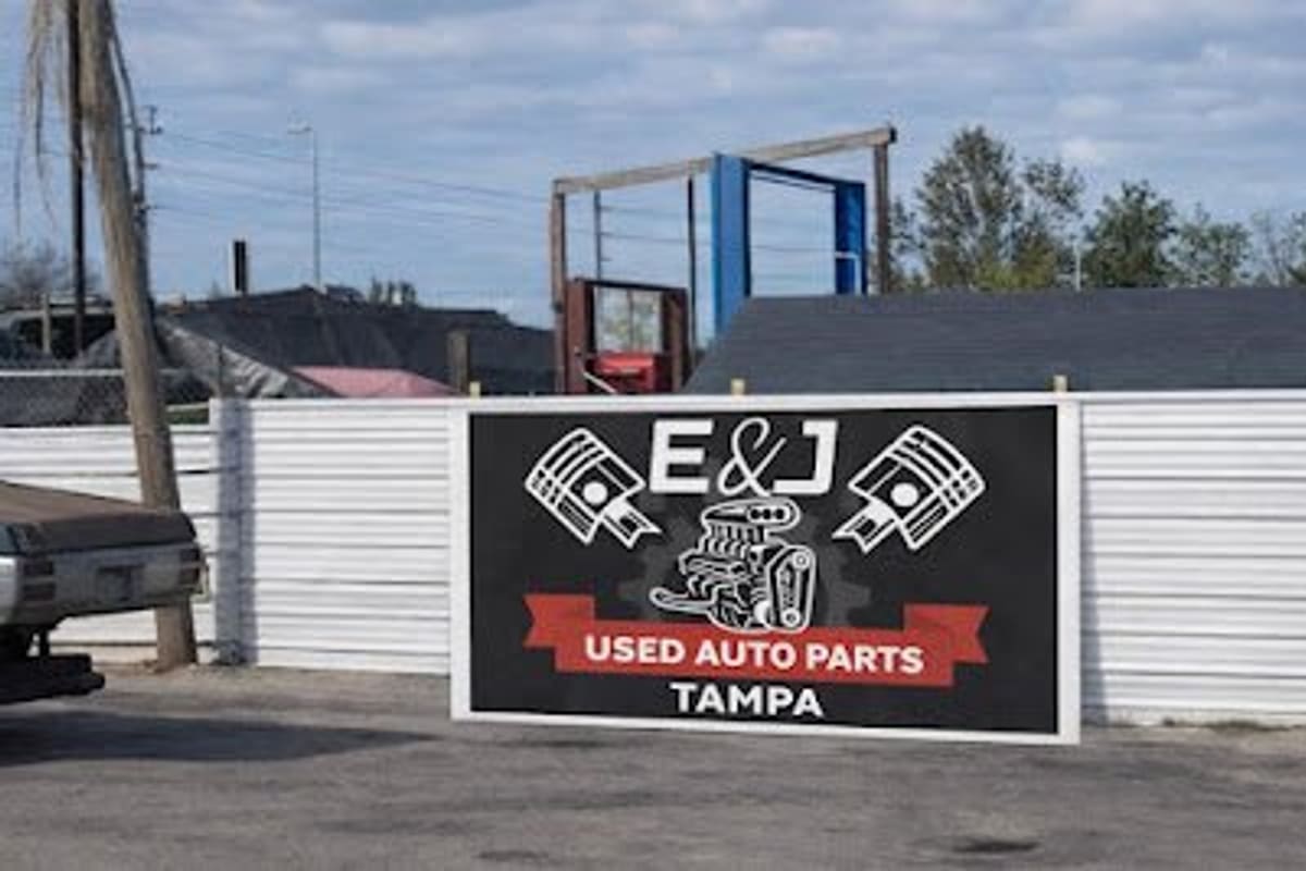 E&J Used Auto Parts Tampa: Large black and white sign reading E&J Used Auto Parts Tampa on a corrugated metal fence at an auto yard