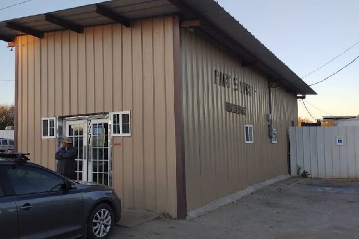 Part S Mart: Tan metal building with a glass entrance door, a sign on the side wall and a gray car parked in front