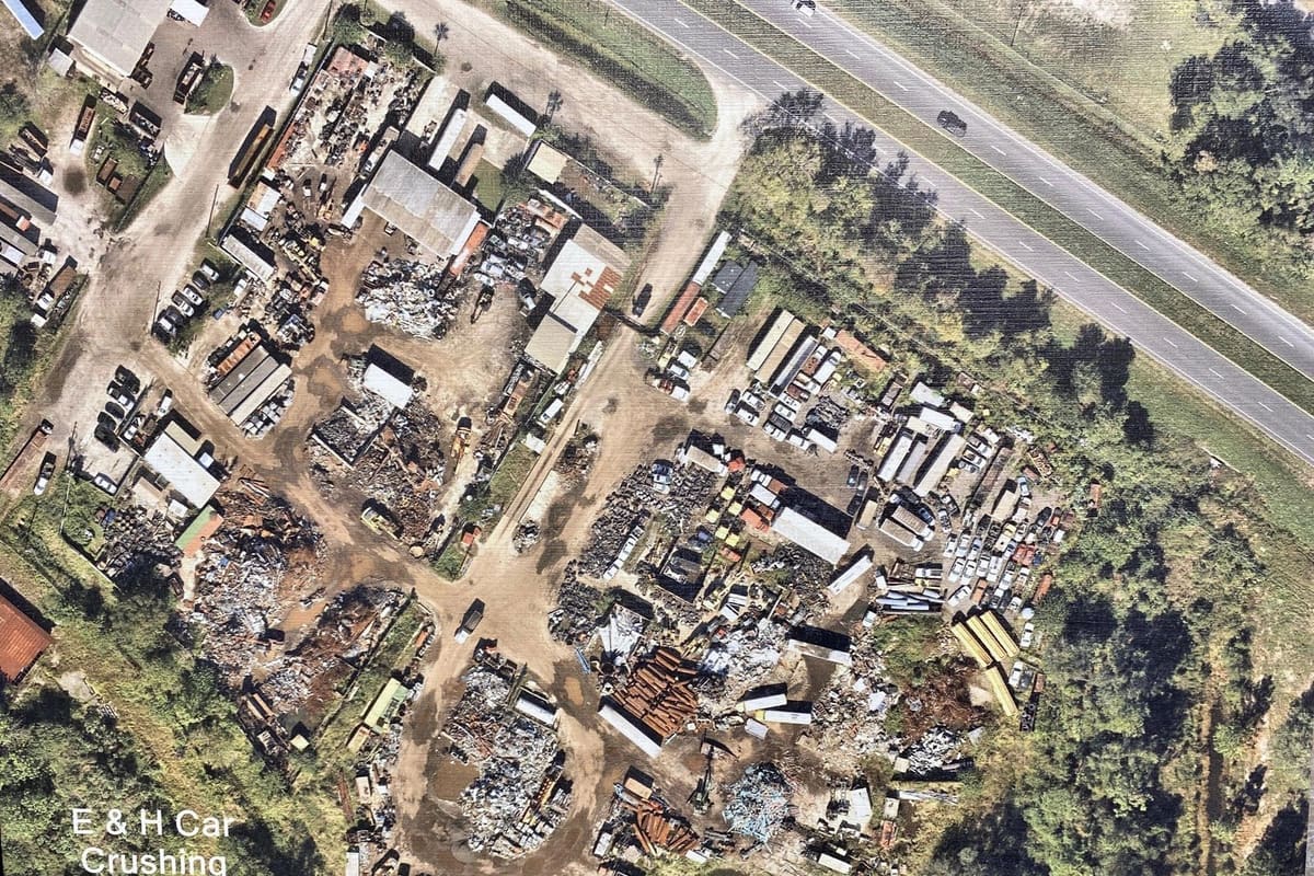 E&H Car Crushing: Aerial view of a salvage yard with piles of scrap metal, rows of vehicles, and buildings beside a highway