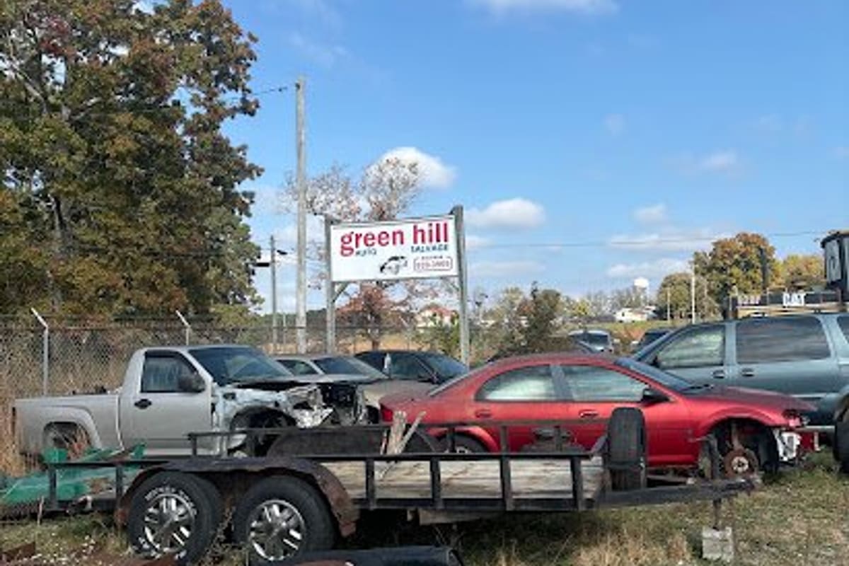 Green Hill Auto Salvage: Salvage yard with wrecked cars, a flatbed trailer and stacked tires on grass beneath a roadside sign and blue sky