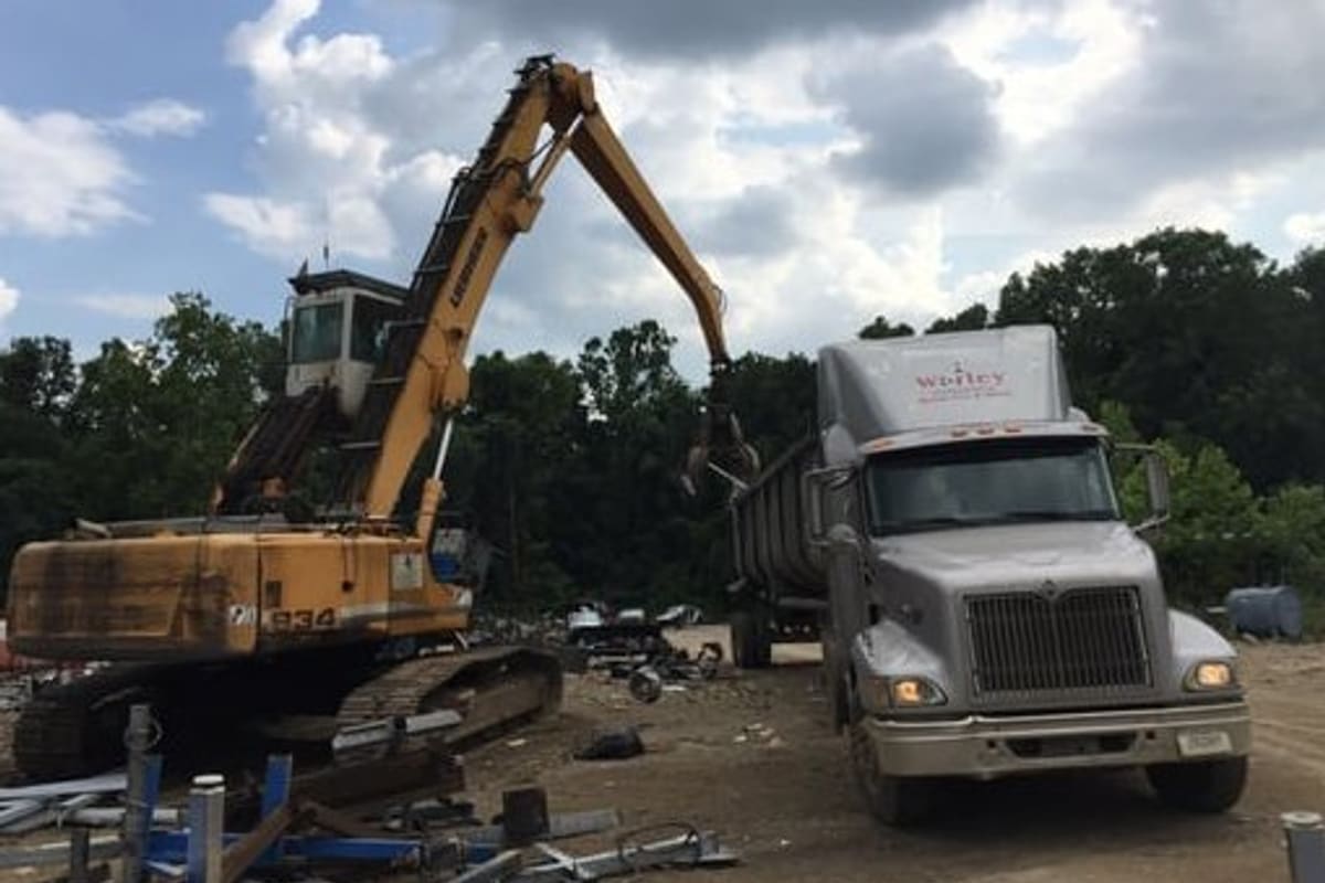 Worley Brothers Scrap Iron & Metal: Yellow material handler loading scrap into a gray semi truck in a dirt scrap yard with trees behind