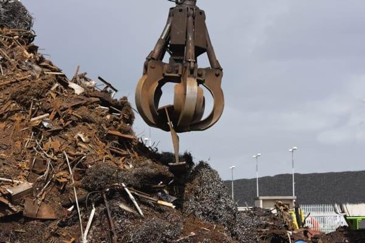 Old Shine Metals: Crane grapple hovering over a large pile of scrap metal in an outdoor salvage yard under a cloudy sky