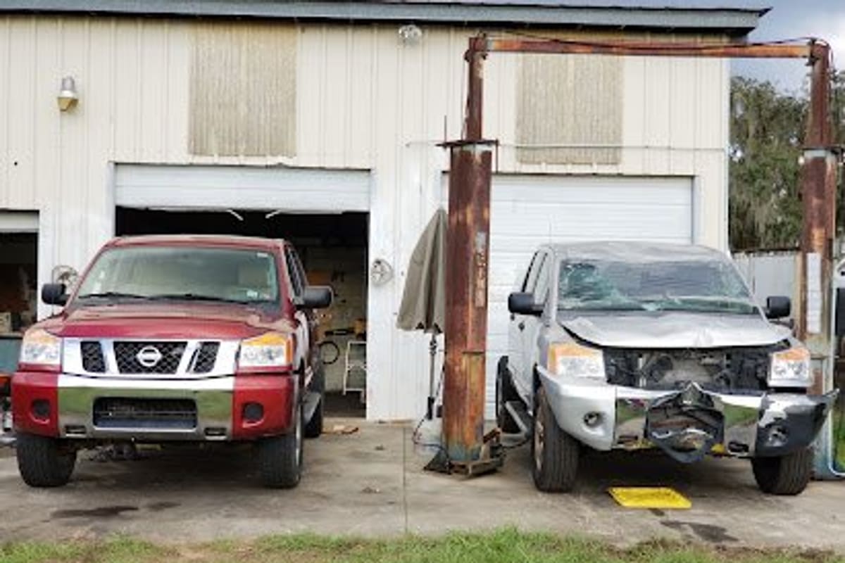 Lake Wales Used Auto Parts: Two Nissan pickup trucks parked in front of a white metal shop building with open garage bays and a rusty lift frame