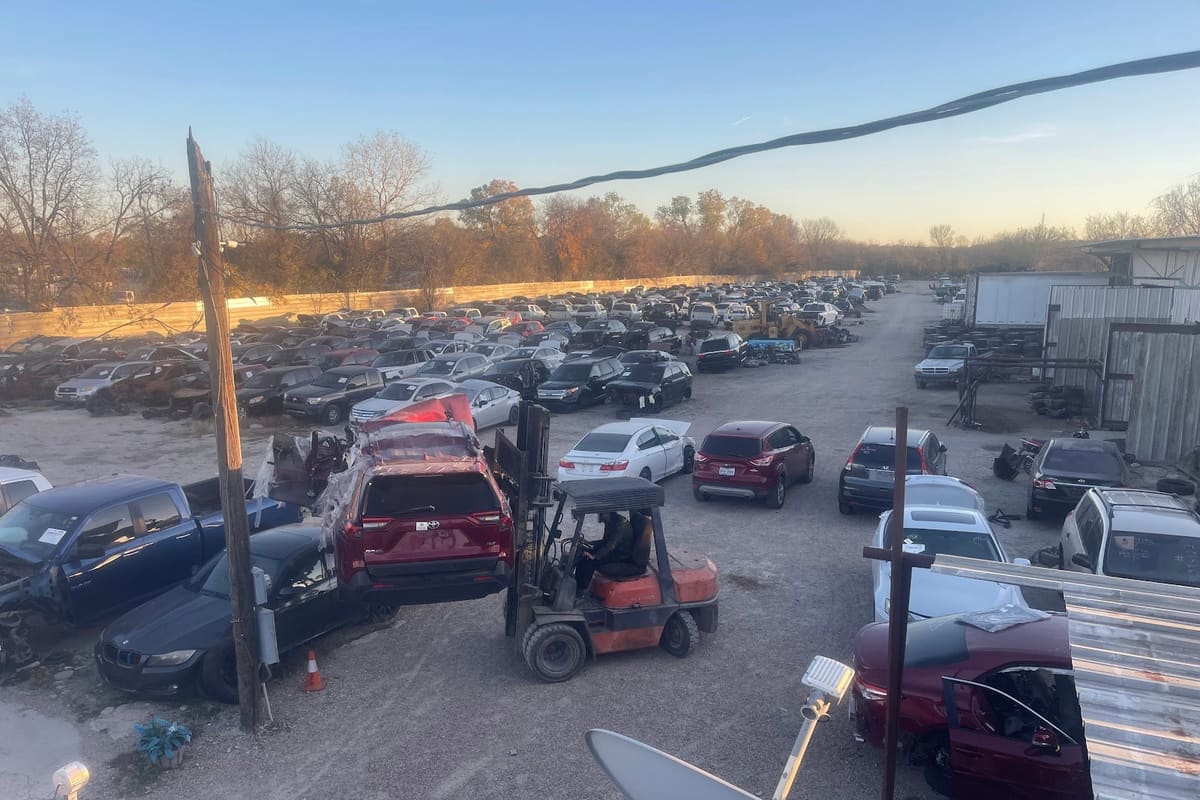 Ace Auto Recycler: Elevated view of a salvage yard with rows of cars and a forklift lifting a red SUV at dusk