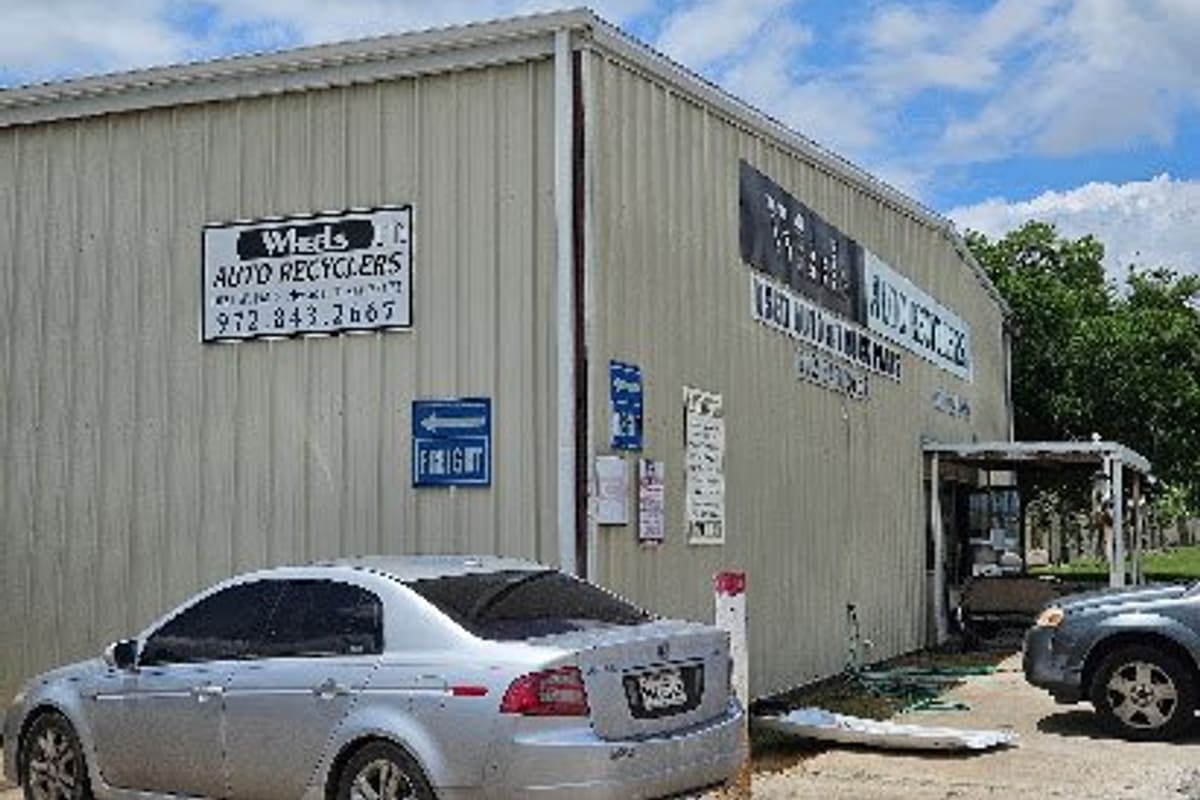 Wheels Auto Recyclers: Tan metal warehouse building with auto recyclers signs, a phone number, and cars parked in front on a gravel lot