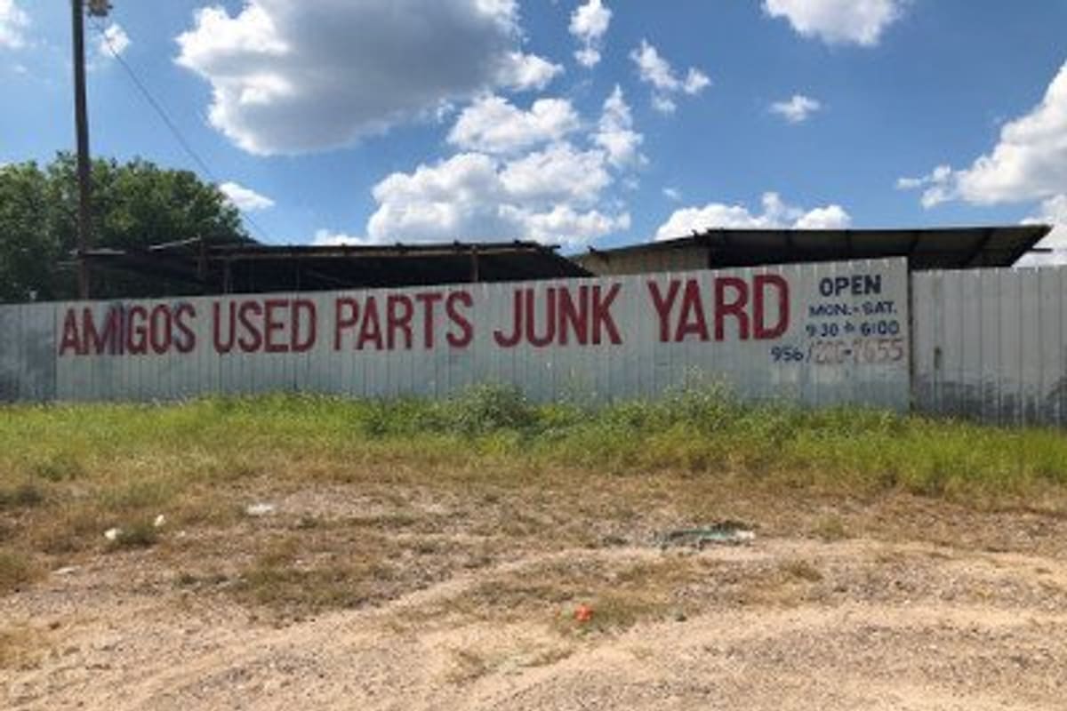Amigos Used Parts Junk Yard: Painted fence sign reading Used Parts Junk Yard with hours and phone number, above a dirt lot and grass under a blue sky