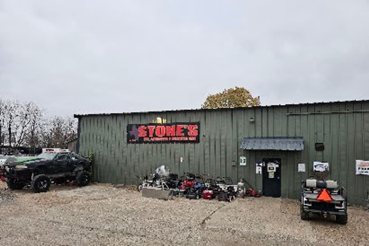 Stone's Wrecking Yard: Green metal shop building with a red sign, a side door, a gravel lot, lawn mowers, a golf cart and a lifted truck
