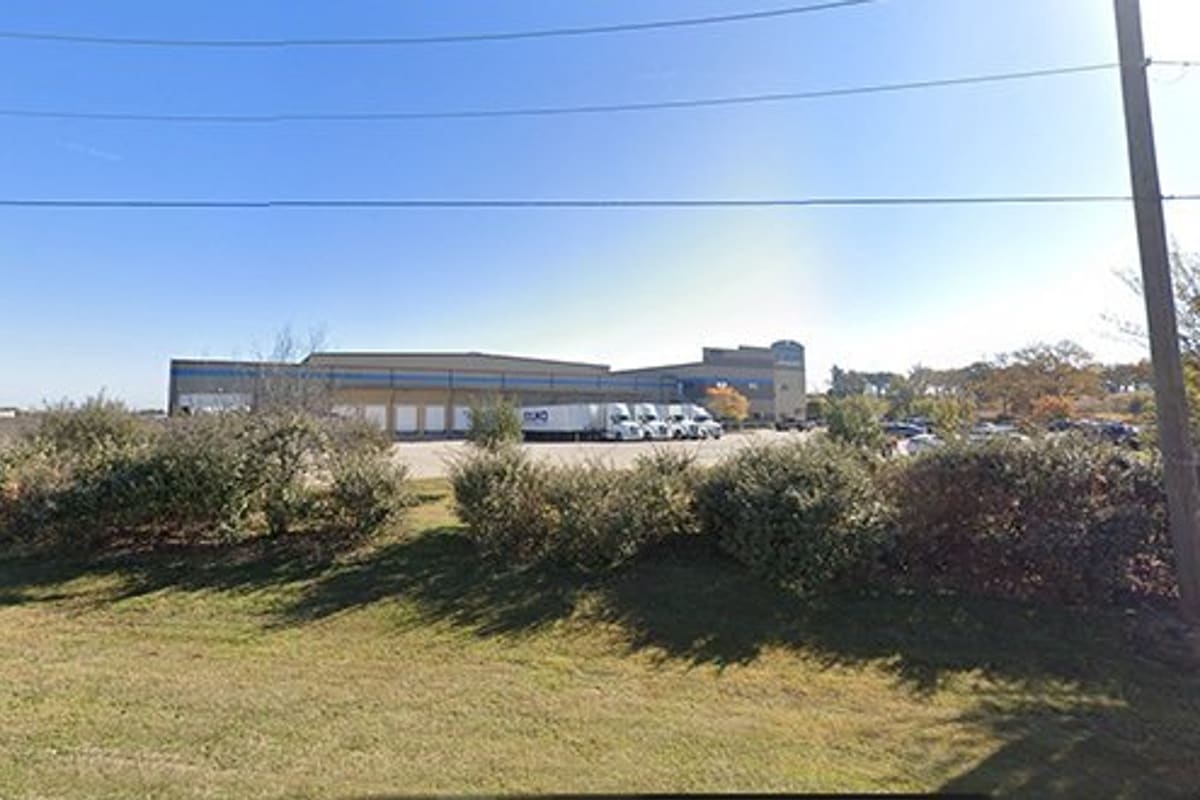 LKQ North Texas - Burelson: Large warehouse building with loading docks and parked semi trucks behind a grassy roadside hedge