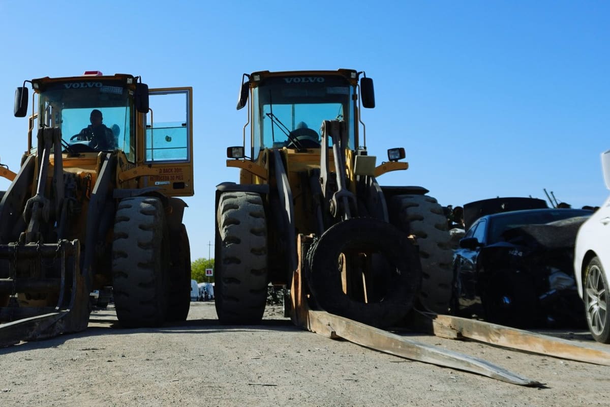 AAA Auto Recyclers: Two yellow wheel loaders with forks parked on a gravel salvage yard, with wrecked cars on either side under a clear blue sky