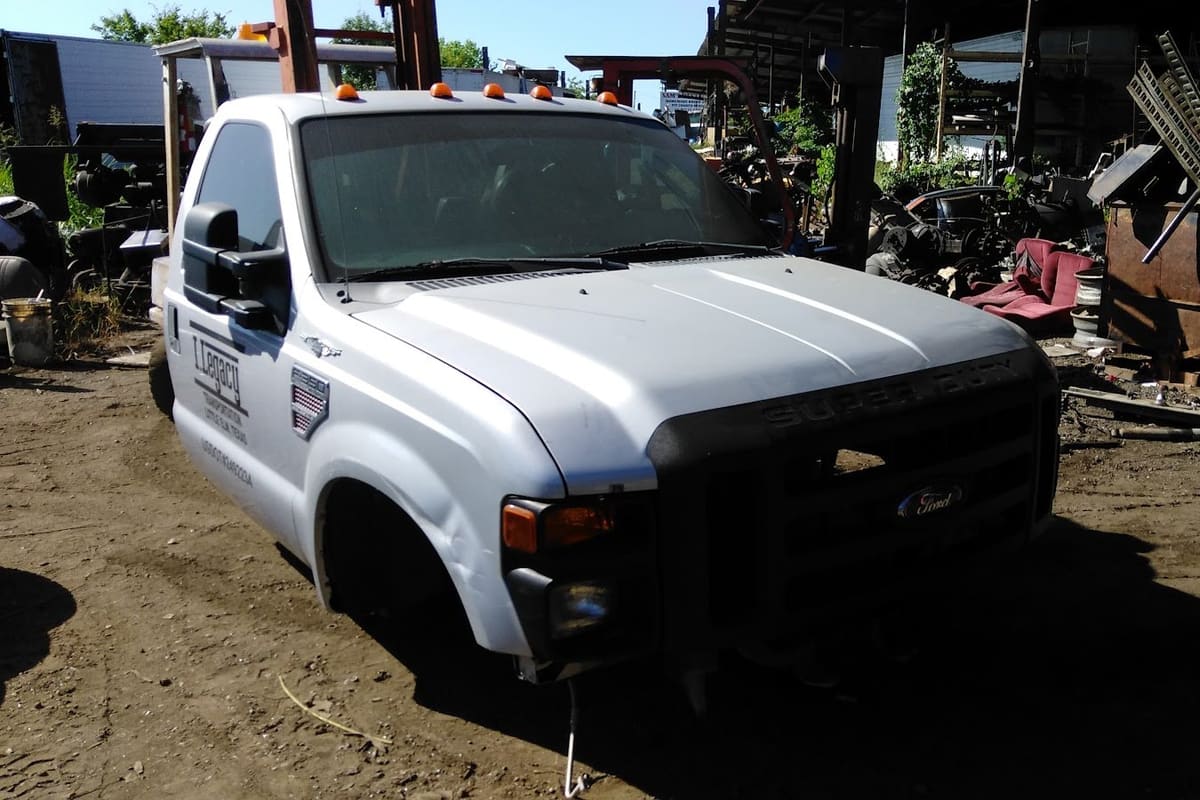 De Leon Truck Parts: White truck cab with no bed sitting in a dirt salvage yard surrounded by scrap parts, bins and covered work areas