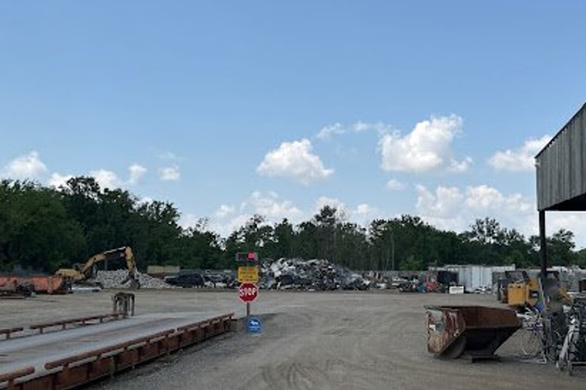 D & D Auto Salvage: Wide daytime view of a gravel salvage yard with a truck scale, excavator, scrap piles and a stop sign under a blue sky