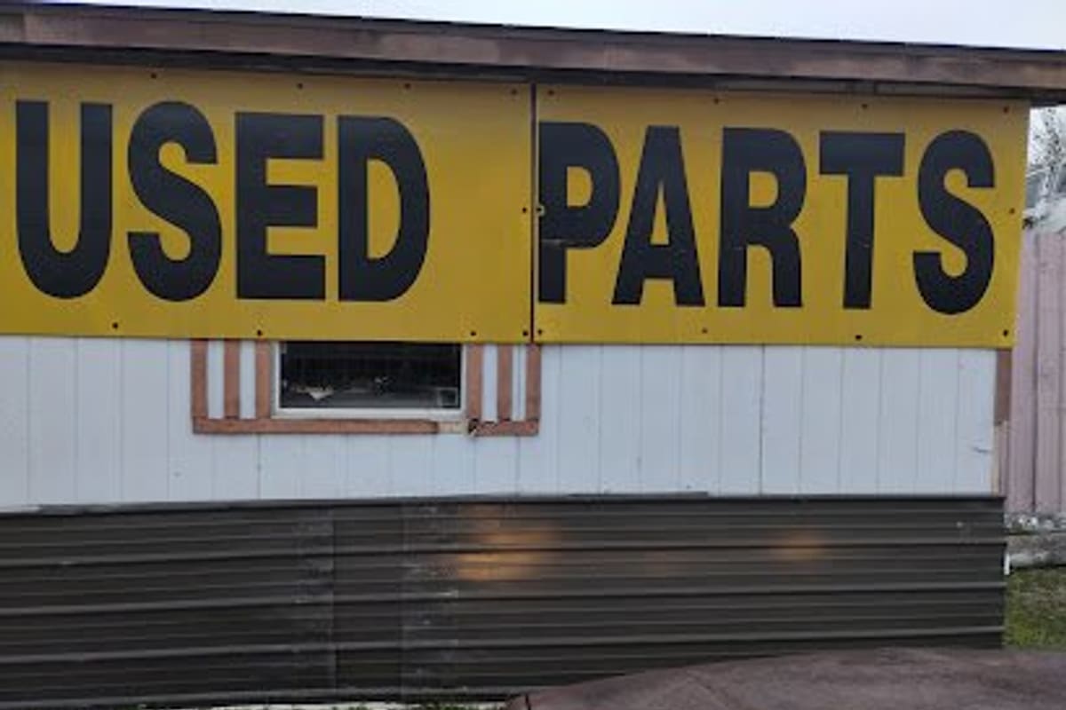 Conroe Auto Recycling Auto & Truck Salvage: Large yellow banner reading USED PARTS on the side of a white and brown metal-skirted building, with a rusty car hood in front