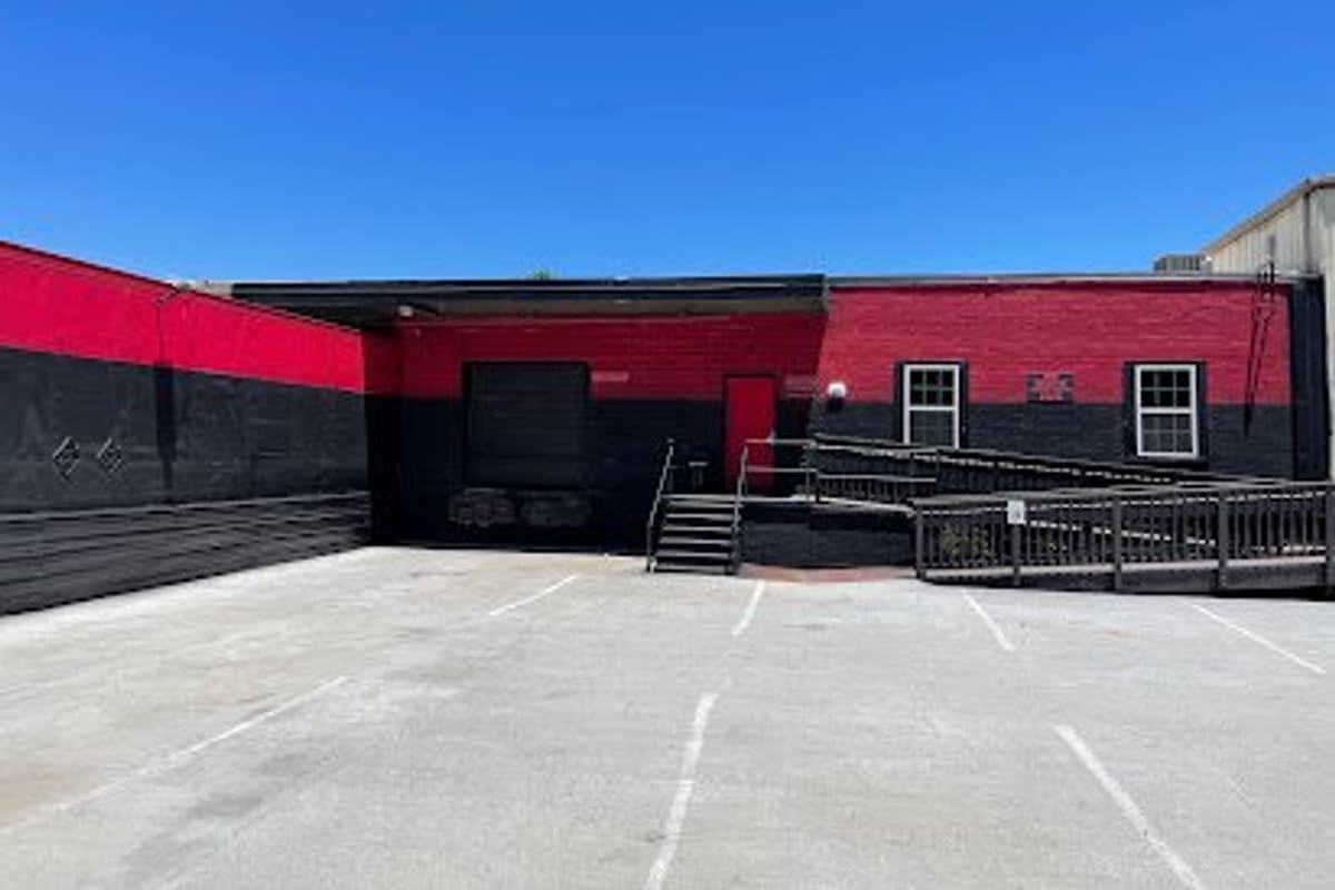 Amk Auto Parts Sales & Service: Red and black brick building with a loading door, entrance stairs and ramp beside an empty paved parking lot under a clear blue sky
