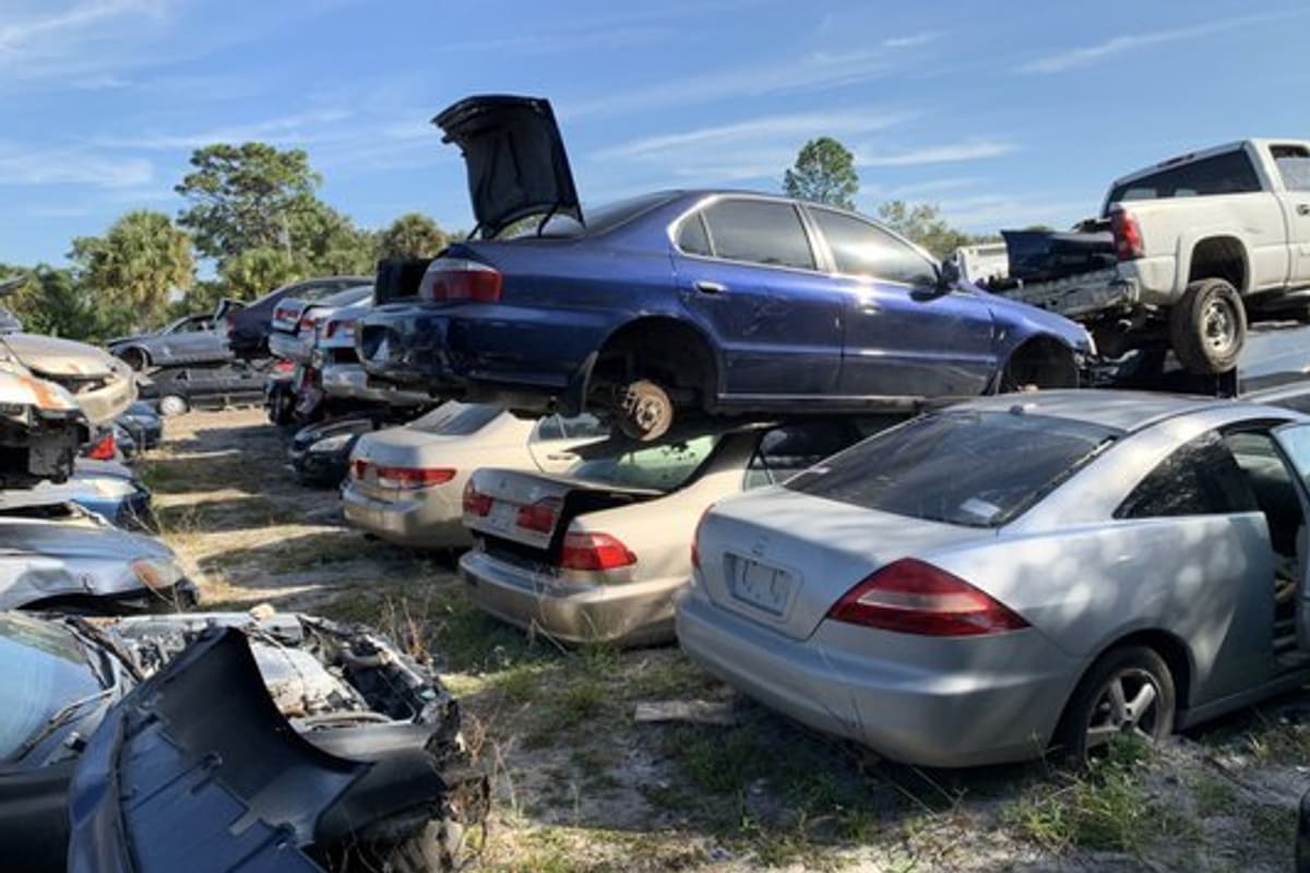 Best Used Auto Parts: Wide daytime view of a salvage yard with wrecked cars parked on grass and stacked, with trees and blue sky behind