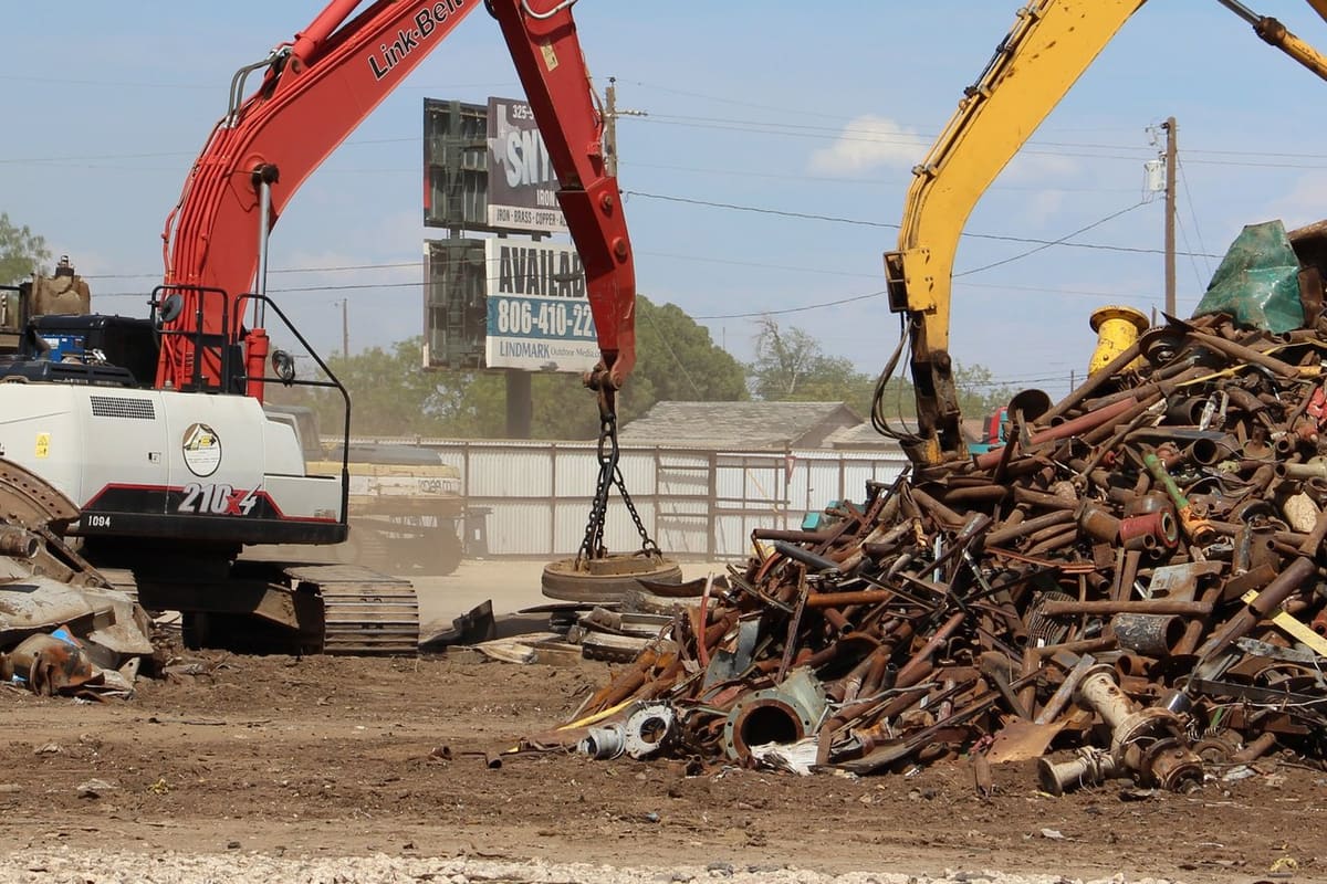 Snyder Iron & Metal: Two excavators with a magnet and grapple sorting a large pile of scrap metal in a dirt yard, with a billboard behind