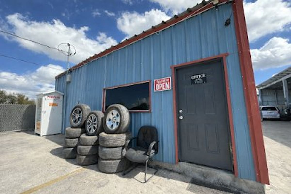 Bee Used Auto Parts: Blue metal office building with a gray door, open sign, and stacked used tires on a paved lot under a partly cloudy sky