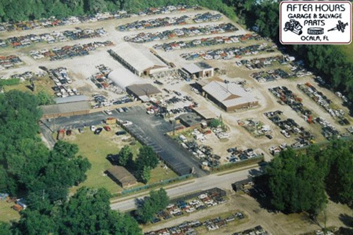 After Hours Salvage: Aerial view of a salvage yard with rows of vehicles, several buildings, and a paved lot surrounded by trees