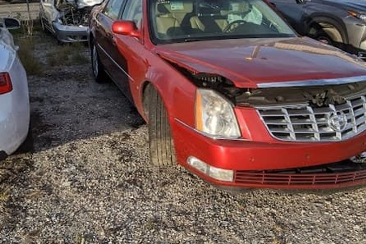 Webbs Salvage: Damaged red sedan parked on gravel in a salvage yard with other wrecked cars and a metal warehouse behind