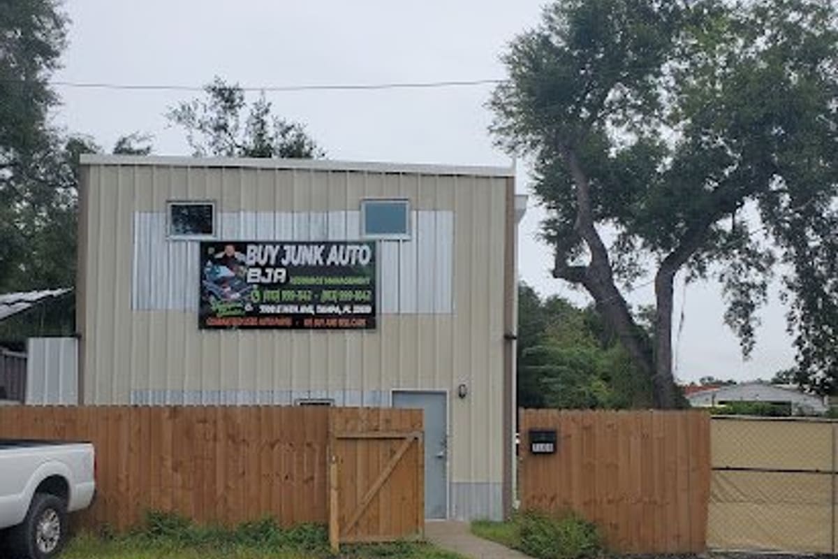 Buy Junk: Two-story tan metal building with a black banner sign, wooden fence and gravel lot in front, under a large oak tree