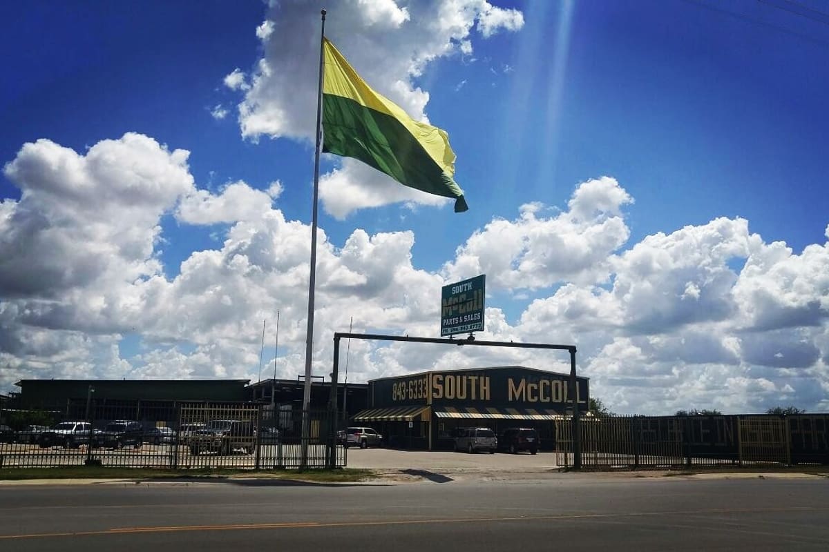 South McColl Used Parts: Roadside salvage yard with a tall flag, sign, fenced lot with vehicles and a building under a blue cloudy sky
