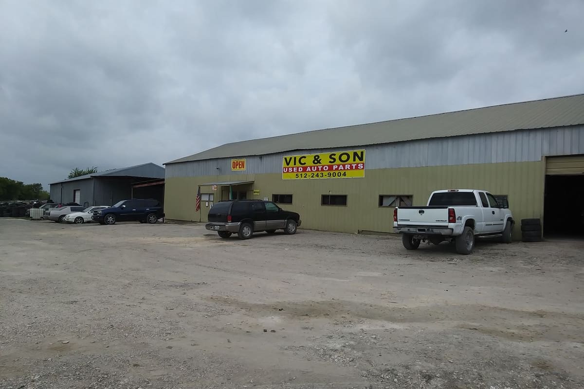 Vic & Son Used Auto Parts: Long tan metal building with a yellow used auto parts sign, gravel lot, and a pickup and SUV parked in front