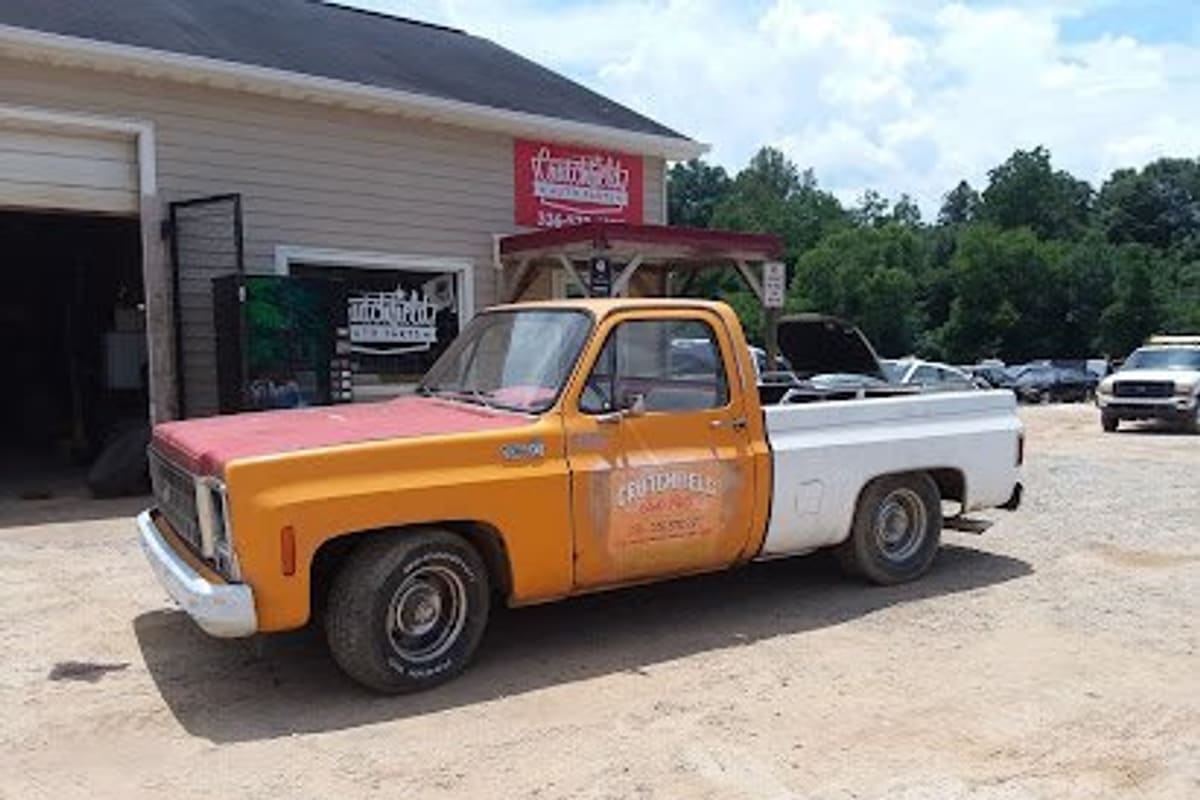 Crutchfield's Auto Parts: Orange and white vintage pickup truck parked on a gravel lot in front of a shop building with a red sign and cars behind