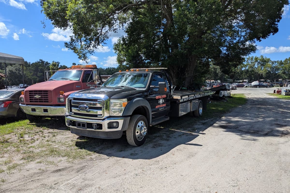 Central Florida Auto Salvage: Black flatbed tow truck and red truck parked on a dirt lane beside a yard of vehicles under large trees