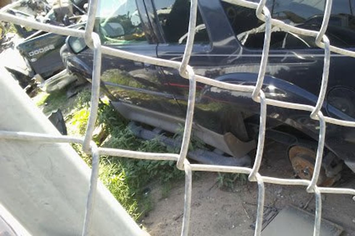 Yonke Amigo Auto Dismantling: Dismantled dark car with a missing wheel seen through a chain-link fence at a salvage yard