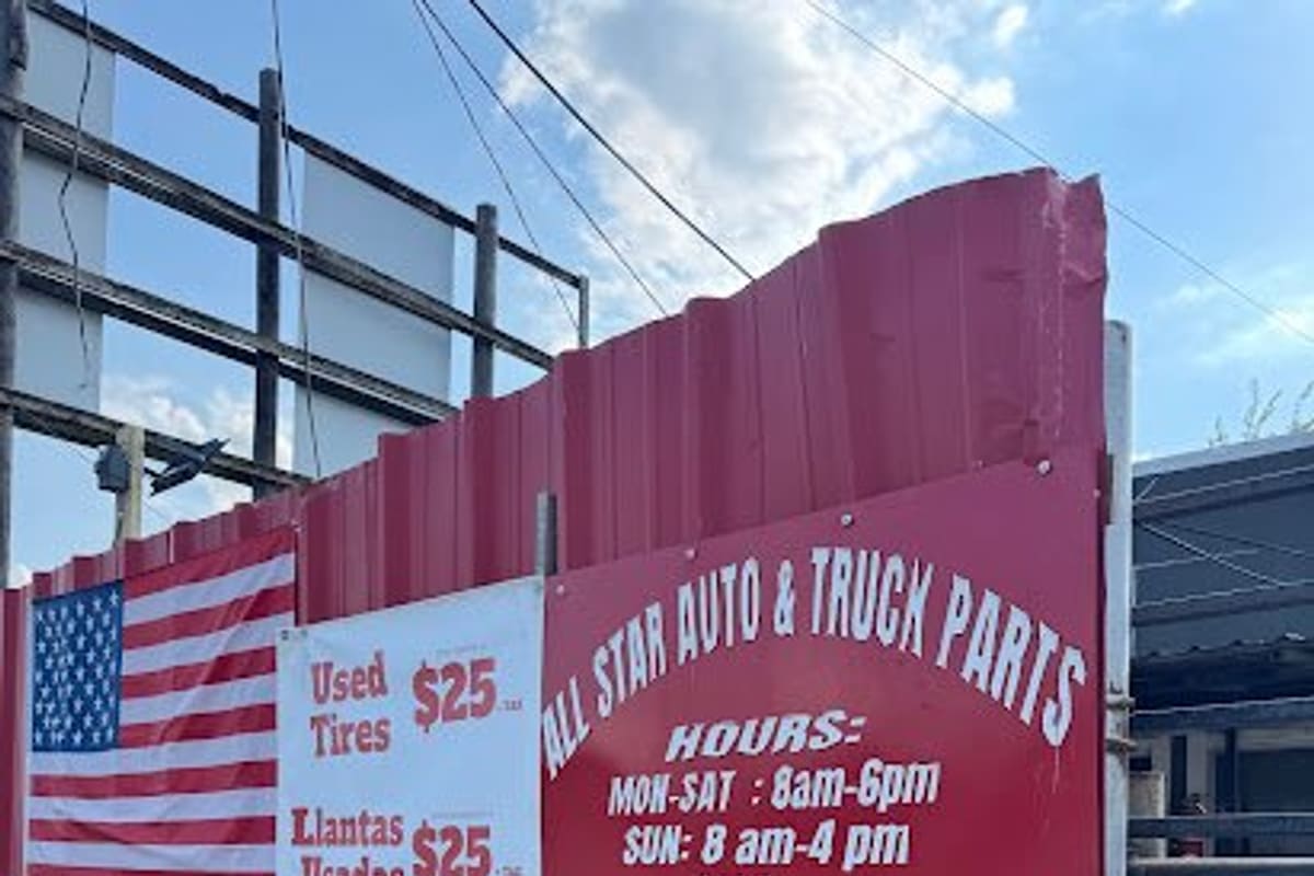 All Star Auto & Truck Parts: Red corrugated metal fence with a hours sign, used tires banner, American flag and a do not block driveway sign