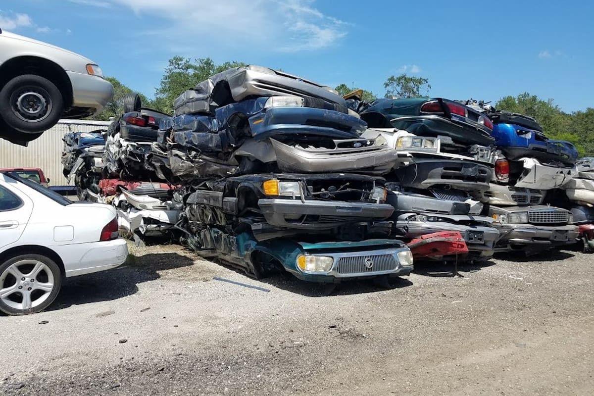 Solutions Auto Parts: Stacks of crushed cars piled high in an outdoor salvage yard on a sunny day, with a white car parked at left