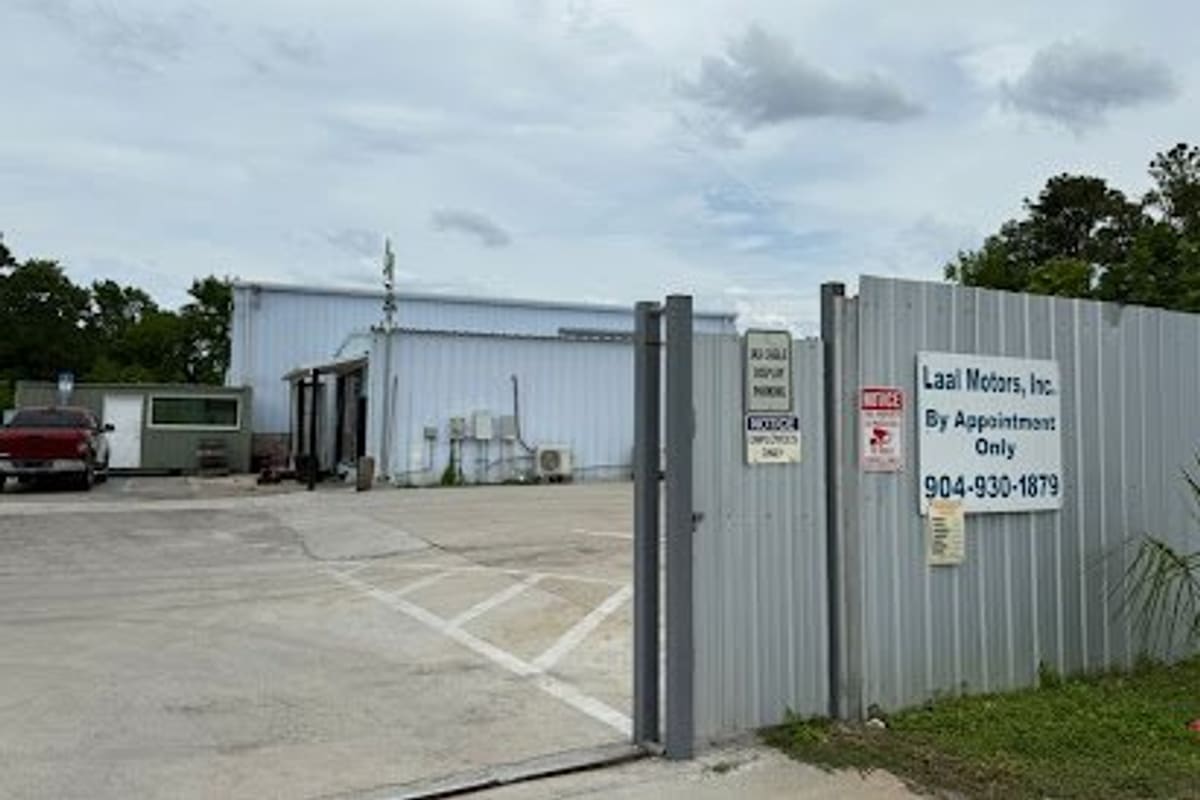 Laalmotors: Open gate beside a corrugated metal fence with a by-appointment sign, leading to a paved lot and metal warehouse building