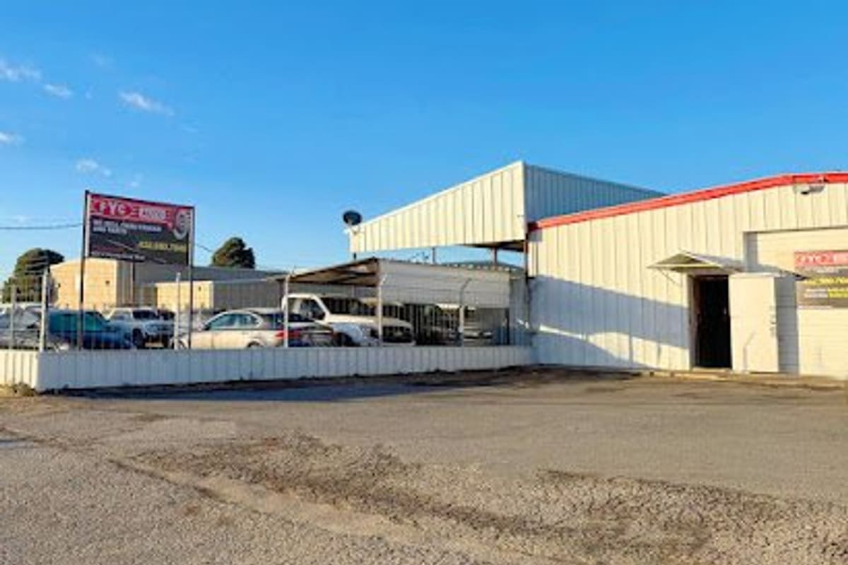 Fyc Auto: White metal shop building with red roofline beside a fenced lot of parked cars and a roadside sign, under a clear blue sky