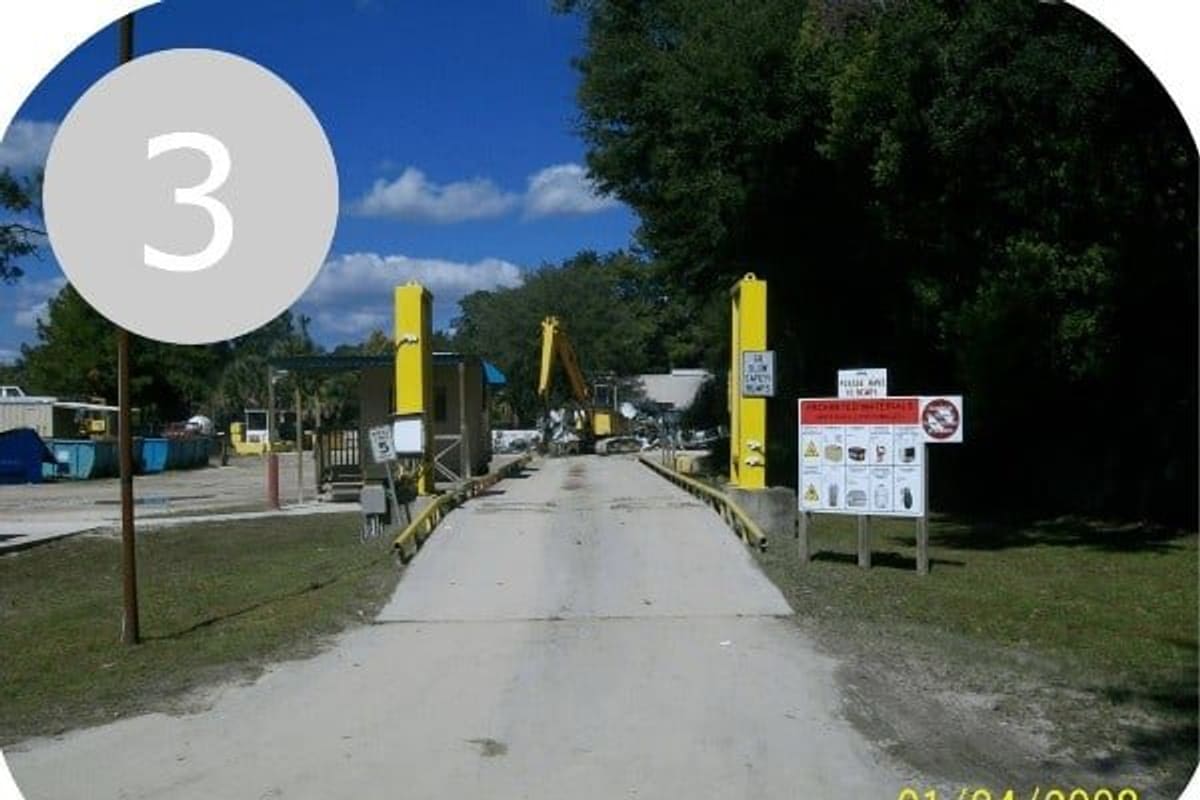 Trademark Metals Recycling: Concrete scale road entering a scrap yard with yellow posts, a small booth, an excavator and a sign of unaccepted materials