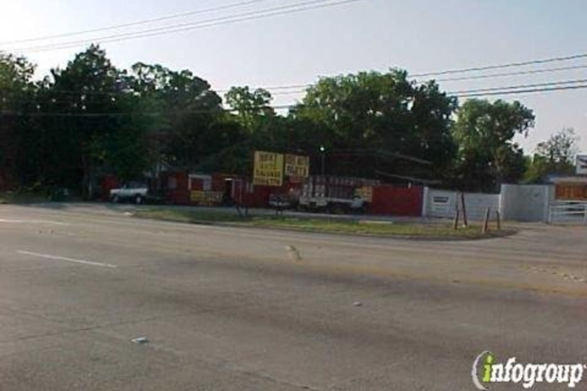 Next Auto Parts: Roadside view of a red salvage yard building with yellow signs, a truck and trees along a wide street