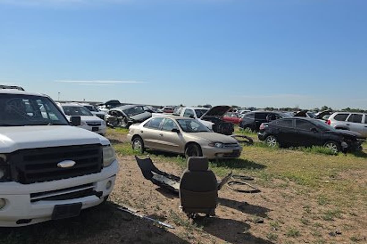 H & H Salvage: Wide daytime view of an open salvage yard with many wrecked cars and a loose car seat on dirt and grass under a blue sky