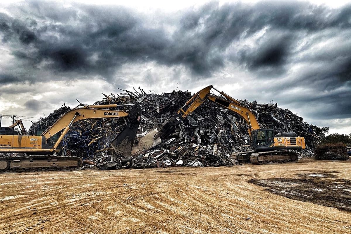 Texas Auto Salvage: Two yellow excavators working at a large pile of scrap metal on a dirt yard under a cloudy sky