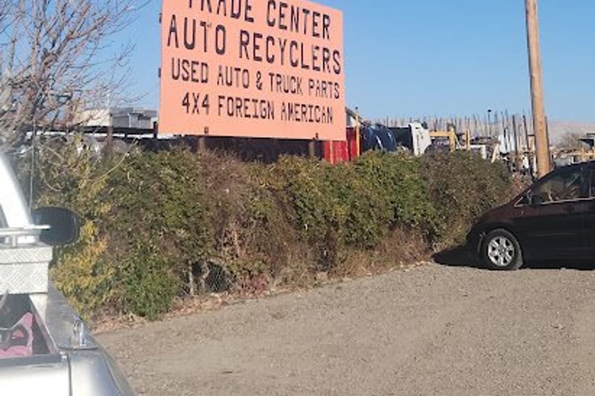 Trade Center Auto Salvage GJ: Large orange roadside sign reading Trade Center Auto Recyclers, used auto and truck parts, above a hedge and gravel lot