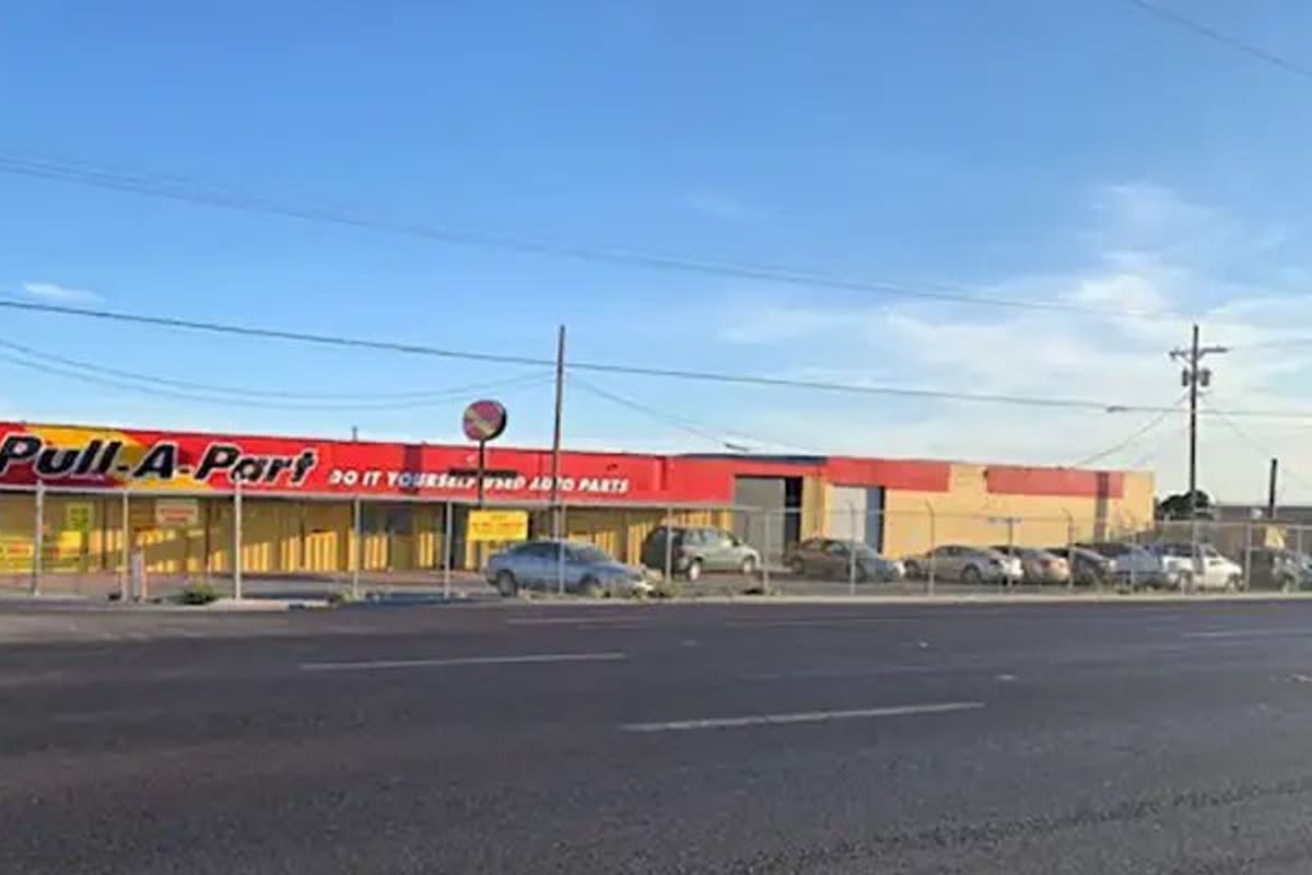 Pull-A-Part El Paso: Long yellow and red self-service auto parts building with a fenced lot of parked cars along a wide road